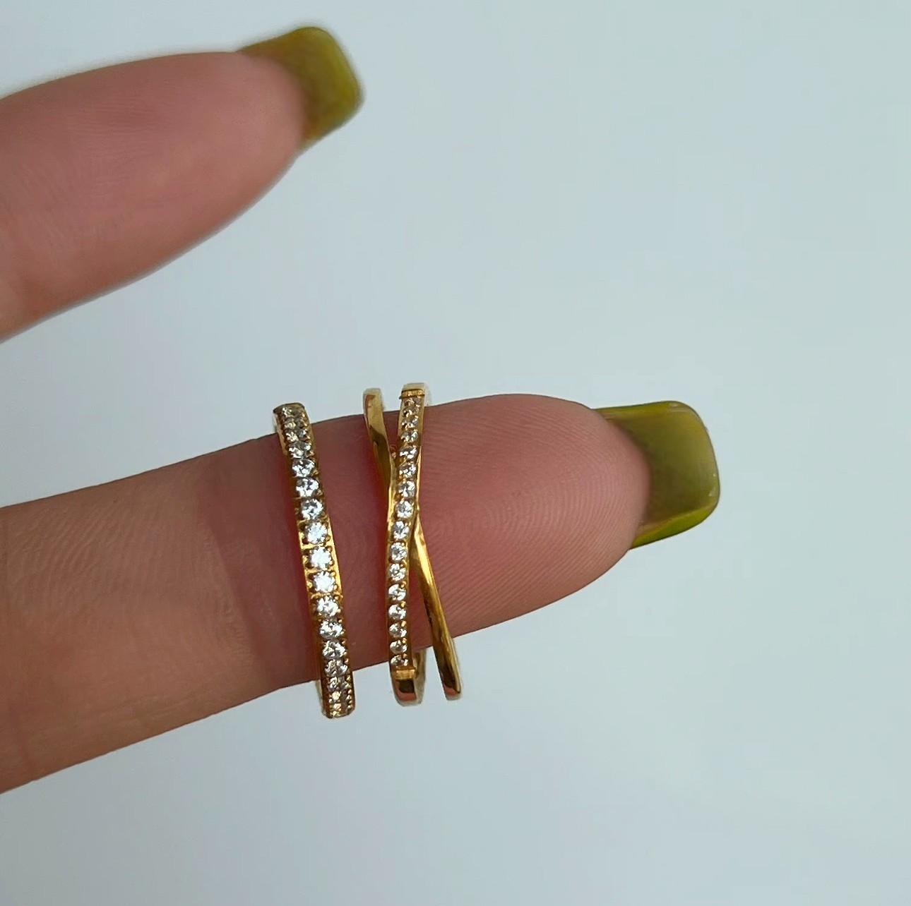 Two gold rings, one band with small clear gemstones and the second with an interlocking design and clear gemstones balanced on a finger. Pictured on a white background