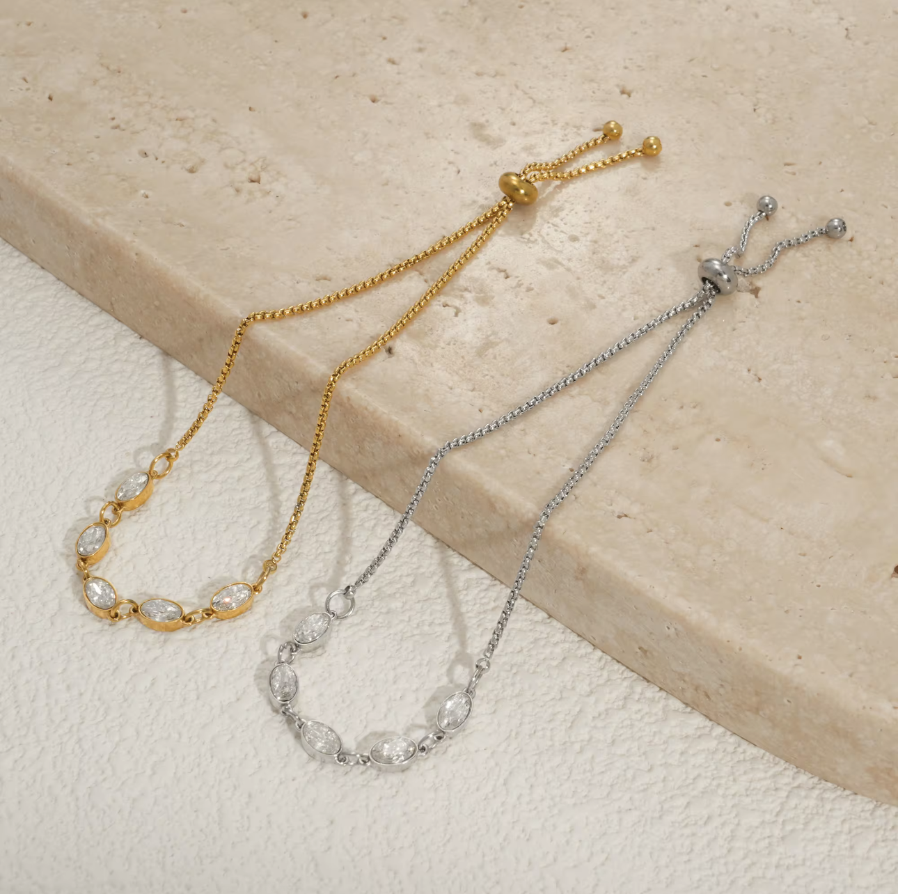 Gold and silver bracelets each featuring five oval shaped clear gemstones on a textured beige surface