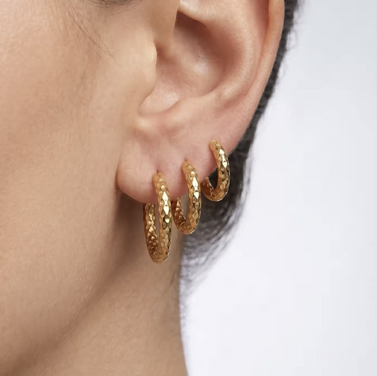 A close-up of a person's ear wearing three textured gold hoop earrings stacked together on the earlobe against a light gray background.