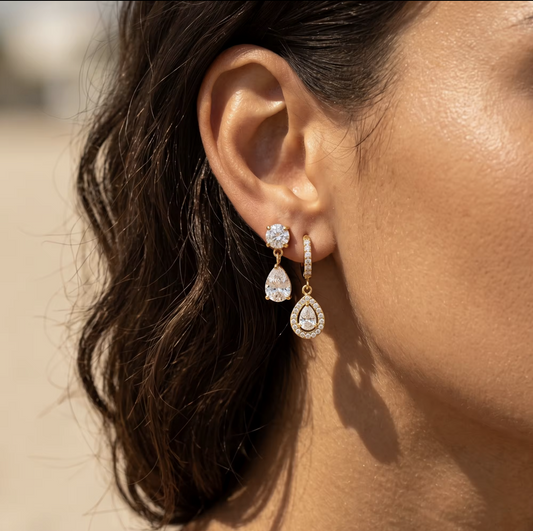 A close-up of a woman's ear wearing a pair of gold drop earrings with clear teardrop-shaped stones and small clear accent stones, photographed in warm natural light.