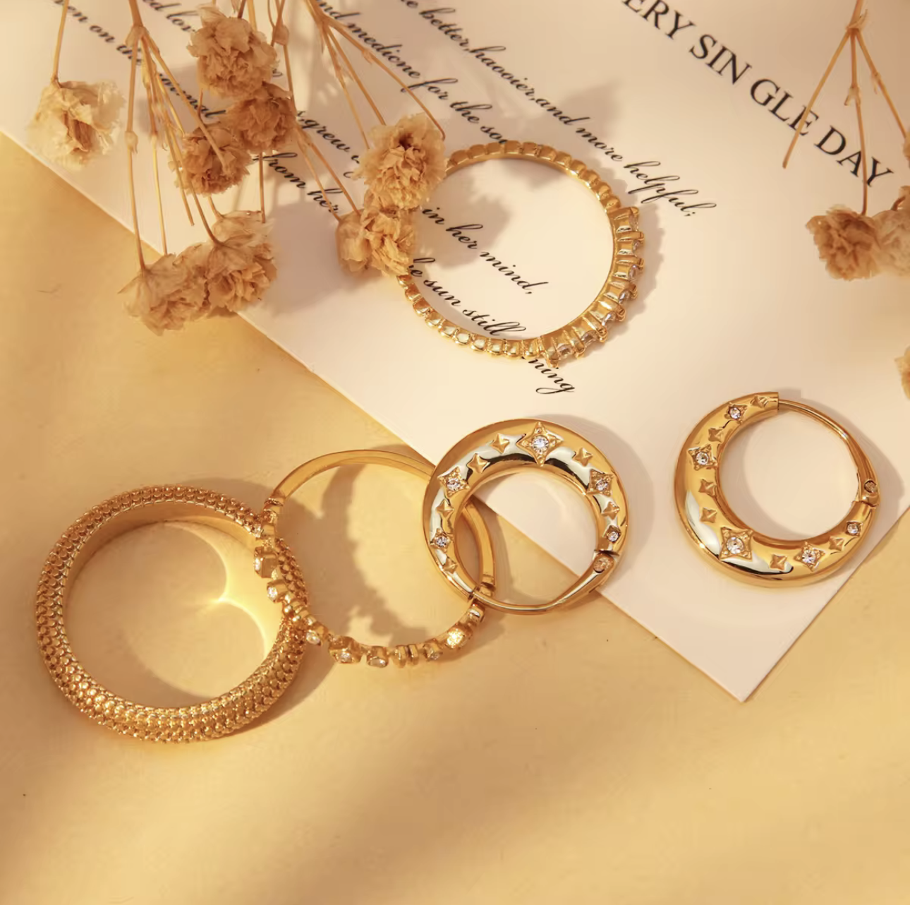 Gold hoop earrings with embedded stones on a textured surface with dried flowers.