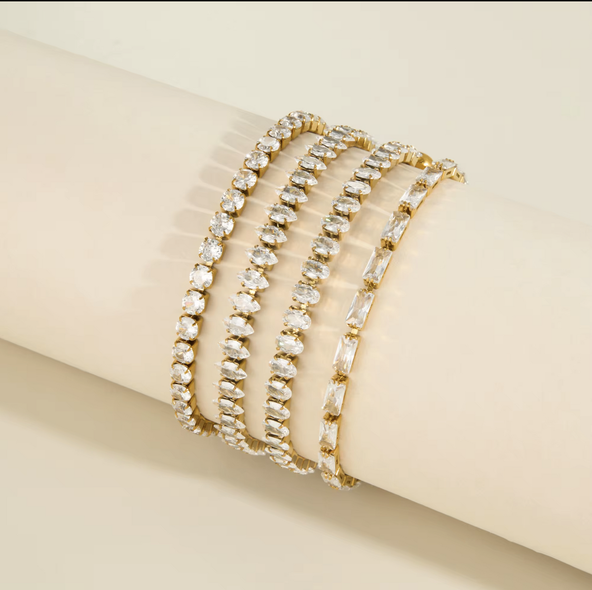 Four gold tennis-style bracelets with clear rhinestones, displayed on a cream-colored cylindrical jewelry stand against a light beige background.