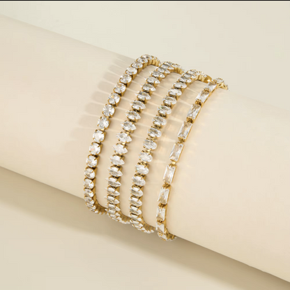 Four gold tennis-style bracelets with clear rhinestones, displayed on a cream-colored cylindrical jewelry stand against a light beige background.