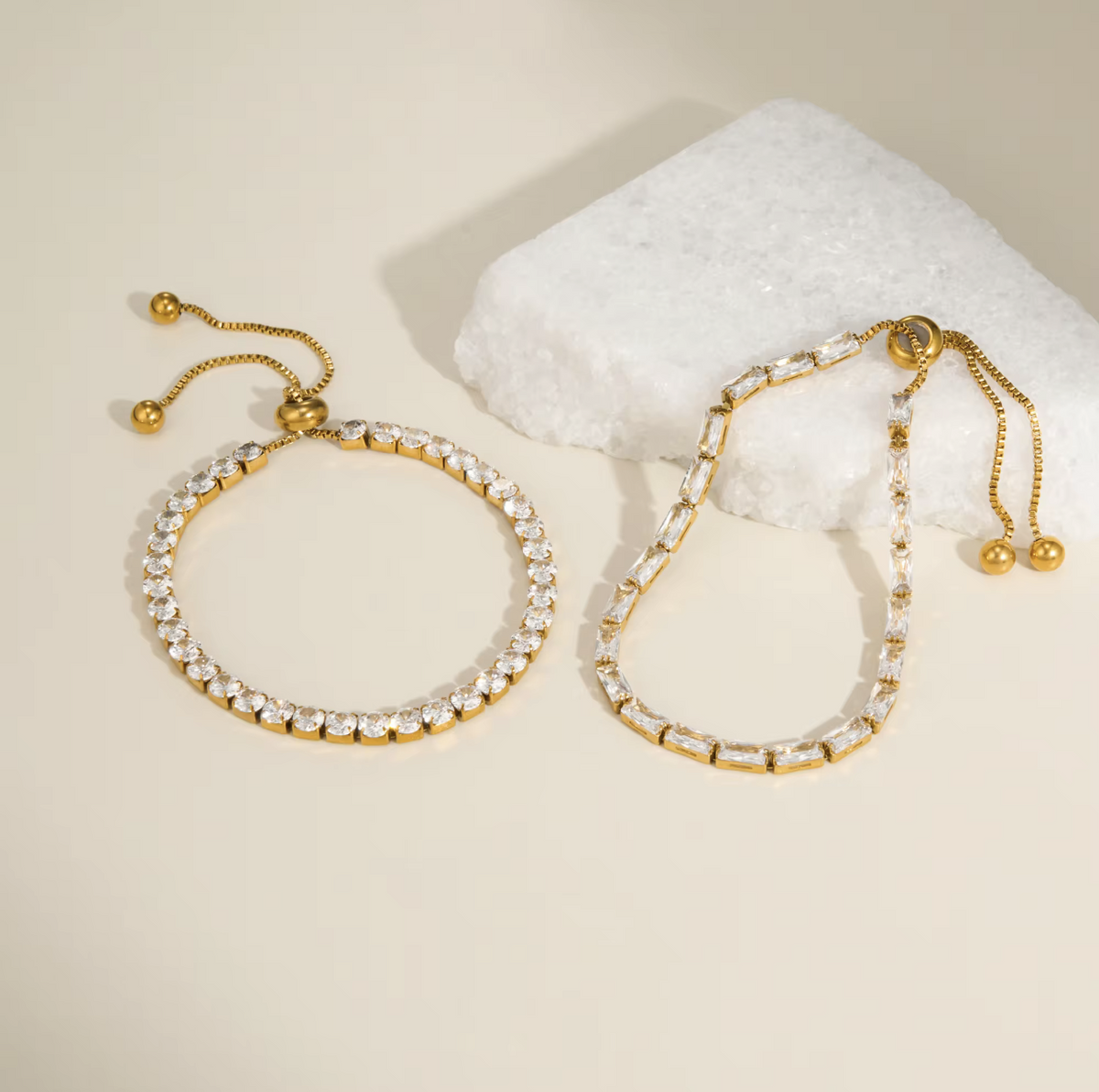 Two gold-toned stretch bracelet-style earrings with clear rhinestone or crystal links, displayed on a warm cream surface beside a white crystal block. No person or real environment is present, so this is Product/Packaging rather than Lifestyle.