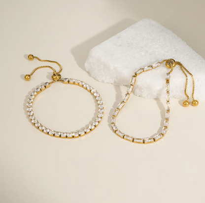 Two gold-toned stretch bracelet-style earrings with clear rhinestone or crystal links, displayed on a warm cream surface beside a white crystal block. No person or real environment is present, so this is Product/Packaging rather than Lifestyle.