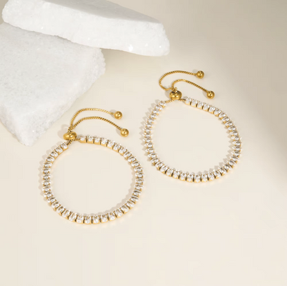 A pair of gold hoop-style earrings with rows of clear rhinestones and adjustable gold chains, photographed on a warm cream background with a white crystal prop.