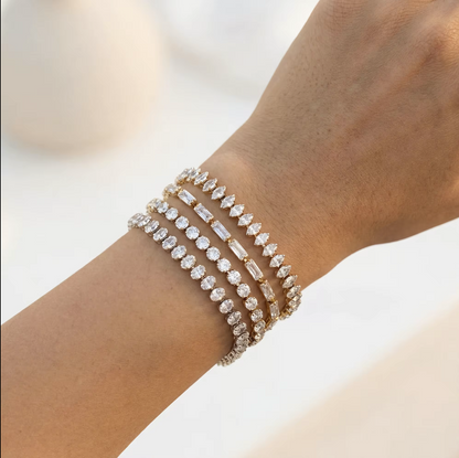 A close-up of a person's wrist wearing four stacked diamond bracelets in silver and gold tones, set against a soft, out-of-focus light background.