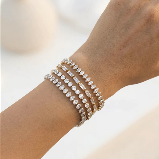 A close-up of a person's wrist wearing four stacked diamond bracelets in silver and gold tones, set against a soft, out-of-focus light background.