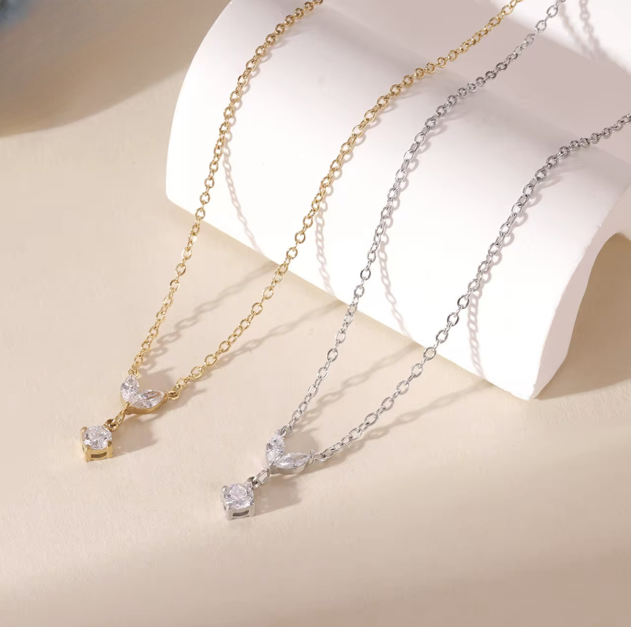 Two necklaces, one gold and one silver. Both with a small marquise and round gemstone pendant on a light beige background
