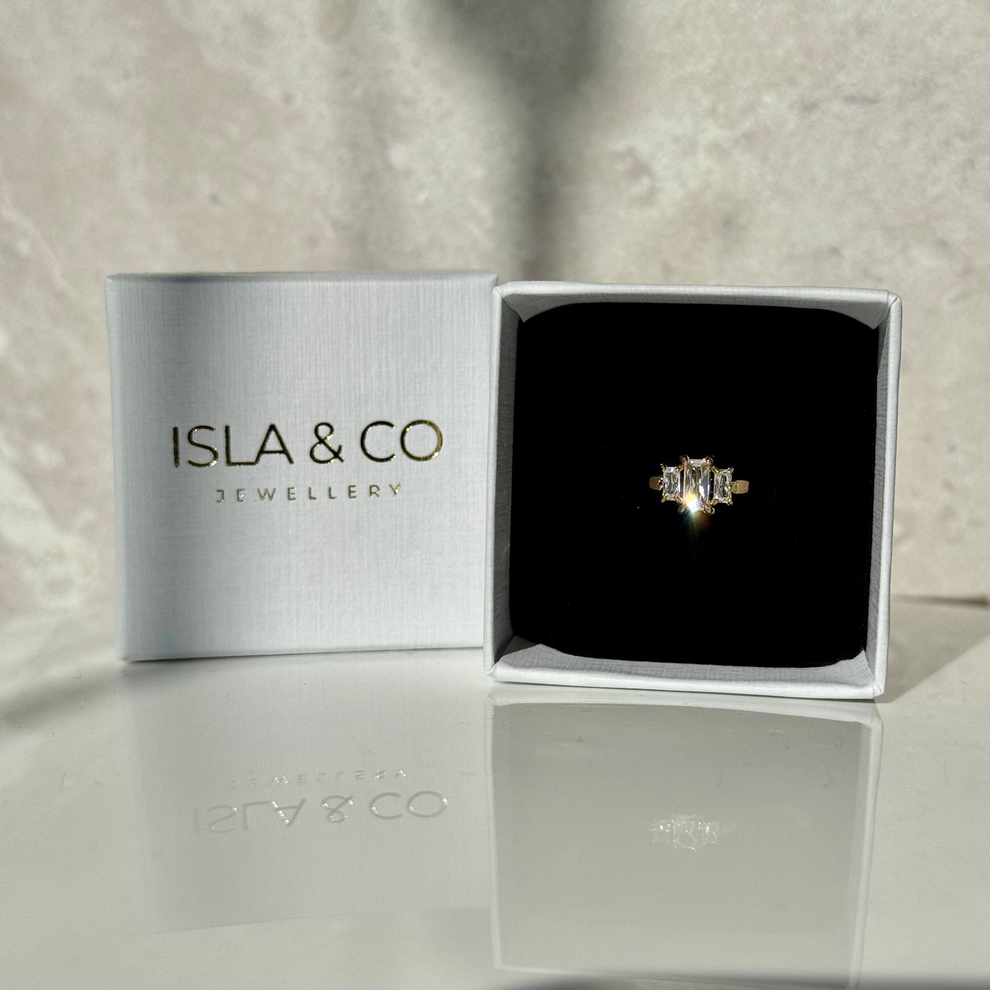 Gold ring with three emerald-cut diamonds. The centre gemstone is larger than the other two either side, which are slightly smaller. Pictured inside a ring box