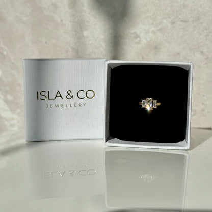 Gold ring with three emerald-cut diamonds. The centre gemstone is larger than the other two either side, which are slightly smaller. Pictured inside a ring box