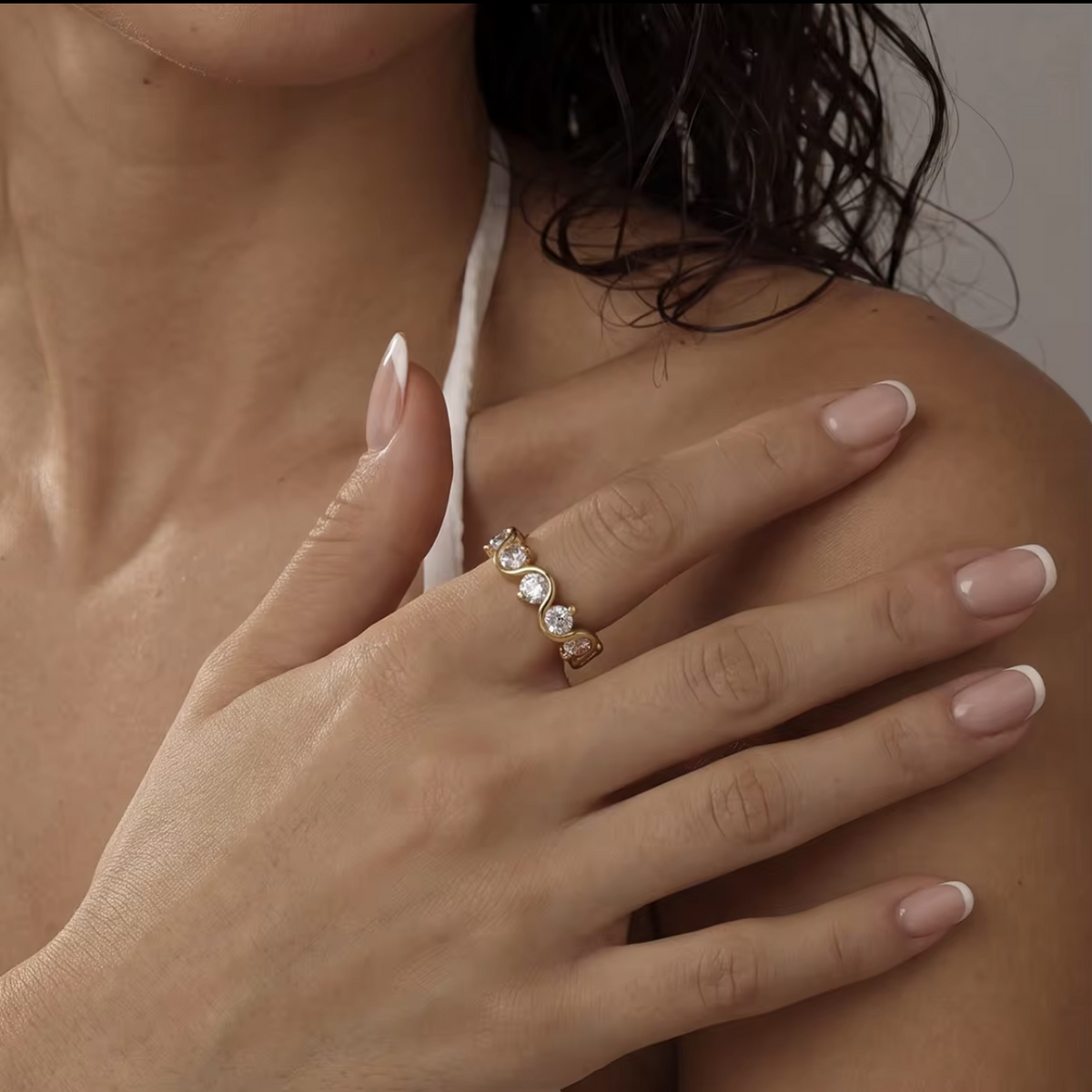 A close-up lifestyle shot of a woman's hand resting on her shoulder, wearing a delicate gold band ring set with four round-cut clear gemstones.