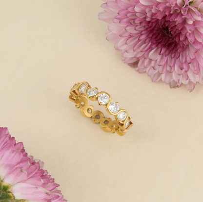 A gold eternity-style ring with five round clear gemstones, photographed on a warm cream background with pink flowers in the corners.