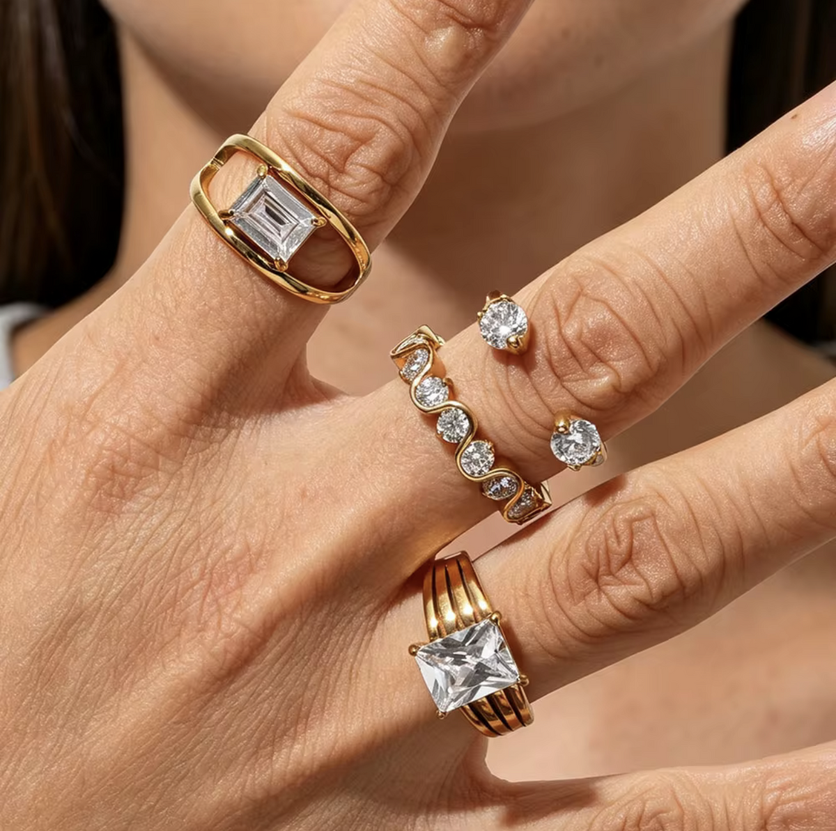 A close-up of a hand wearing multiple gold rings with clear gemstones, including a large rectangular emerald-cut ring, a row of small round-cut stones, and a cluster of three round-cut stones.