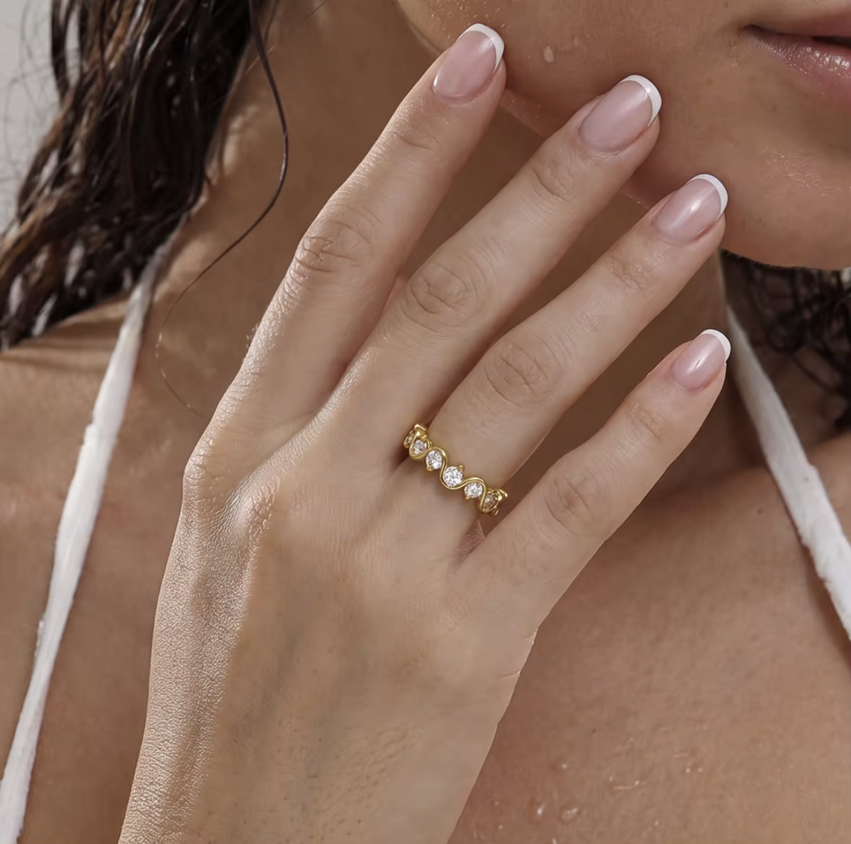 A close-up of a woman's hand wearing a delicate gold ring with five small white pearls, set against a soft-focus background of her face and chest.