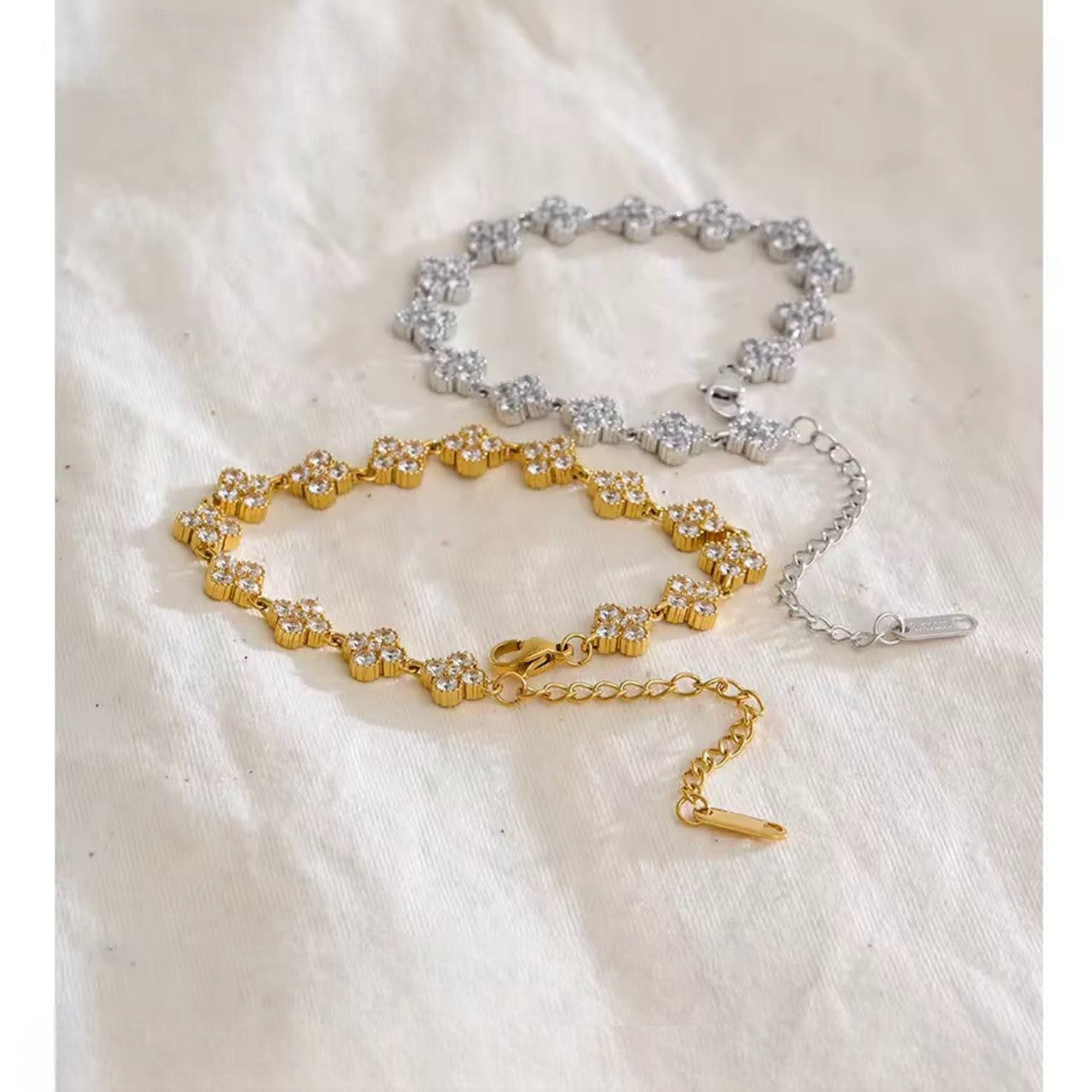 Silver bracelet and gold bracelet, both with clear stones arranged in the shape of a four leaf clover on a biege background