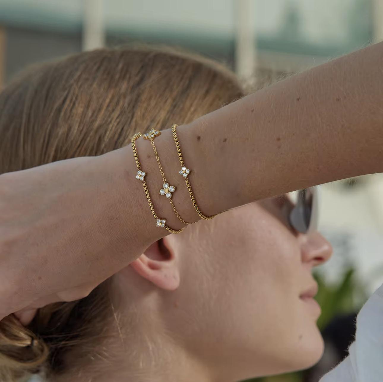 Close-up of a person wearing multiple gold bracelets with floral designs on a blurred outdoor background.