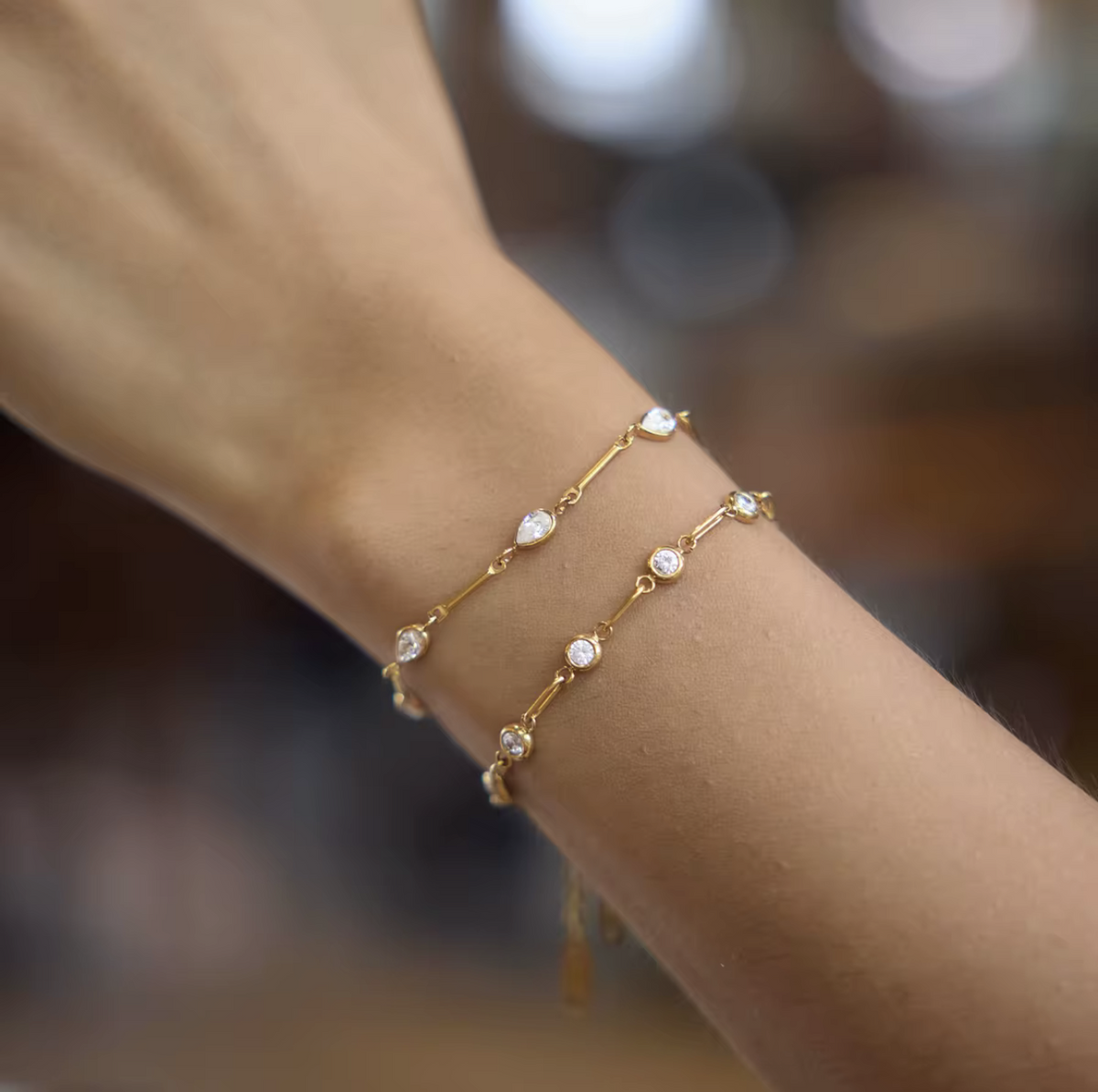 A close-up of a person's wrist wearing two delicate gold bracelets, each adorned with small clear gemstones, against a softly blurred warm brown background.