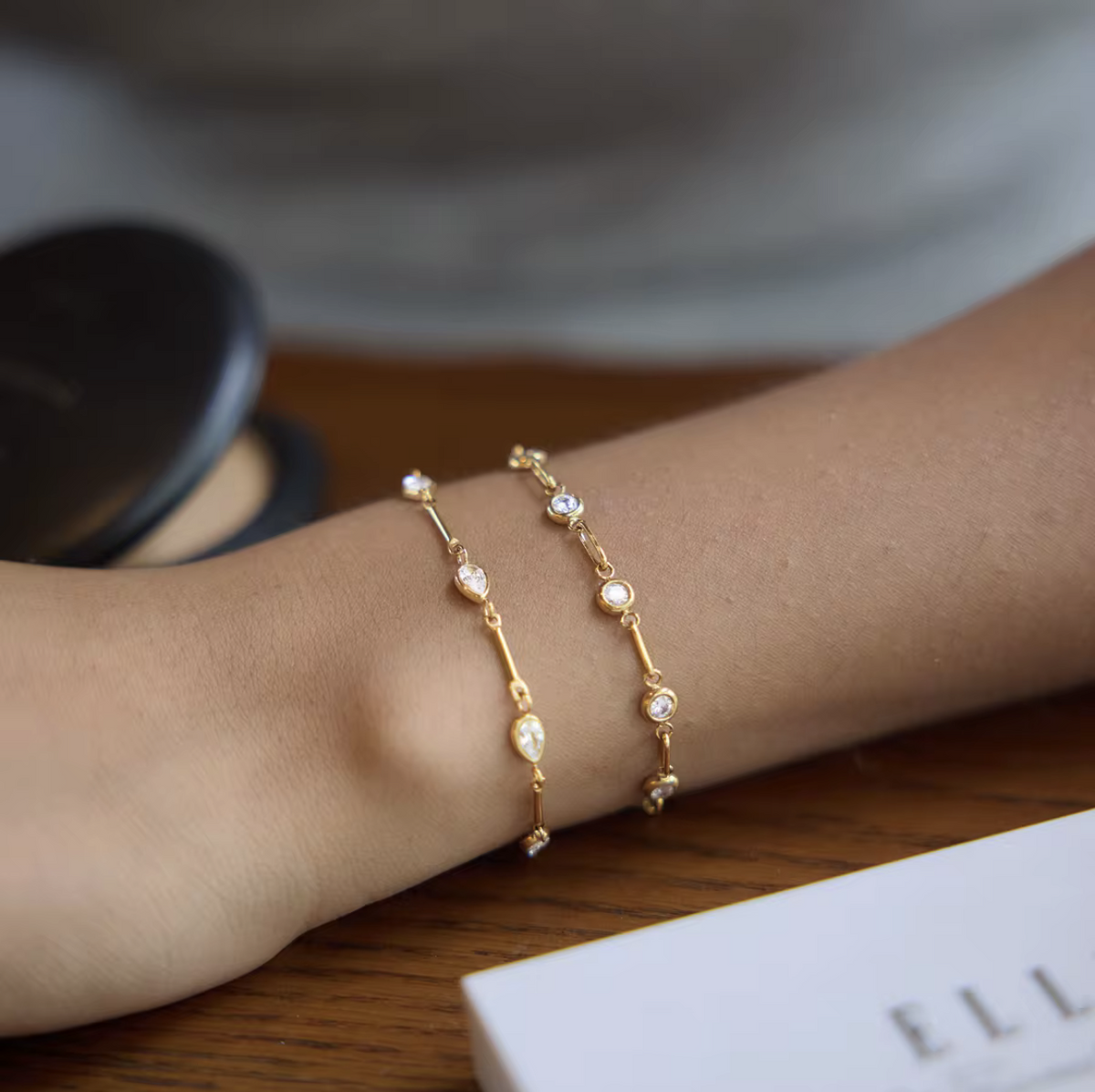 Two delicate gold chain bracelets with small clear stones and pearl-like beads, worn on a person's wrist against a softly blurred indoor background.