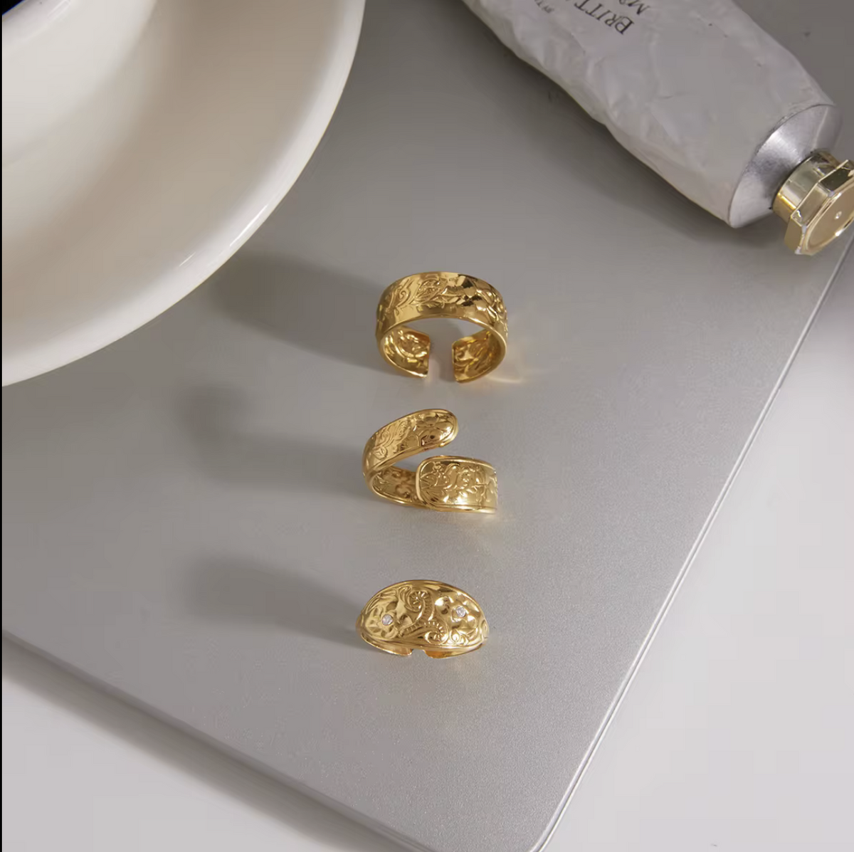 Three textured gold open cuff rings arranged vertically on a silver laptop lid, with a white bowl and a tube of cream partially visible in the background.