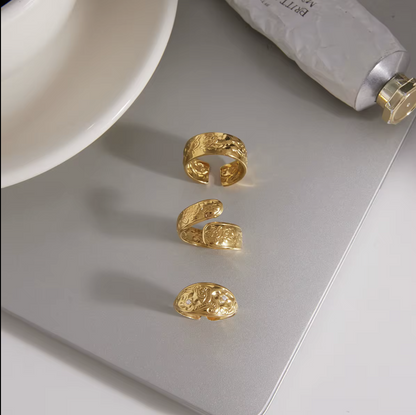 Three textured gold open cuff rings arranged vertically on a silver laptop lid, with a white bowl and a tube of cream partially visible in the background.