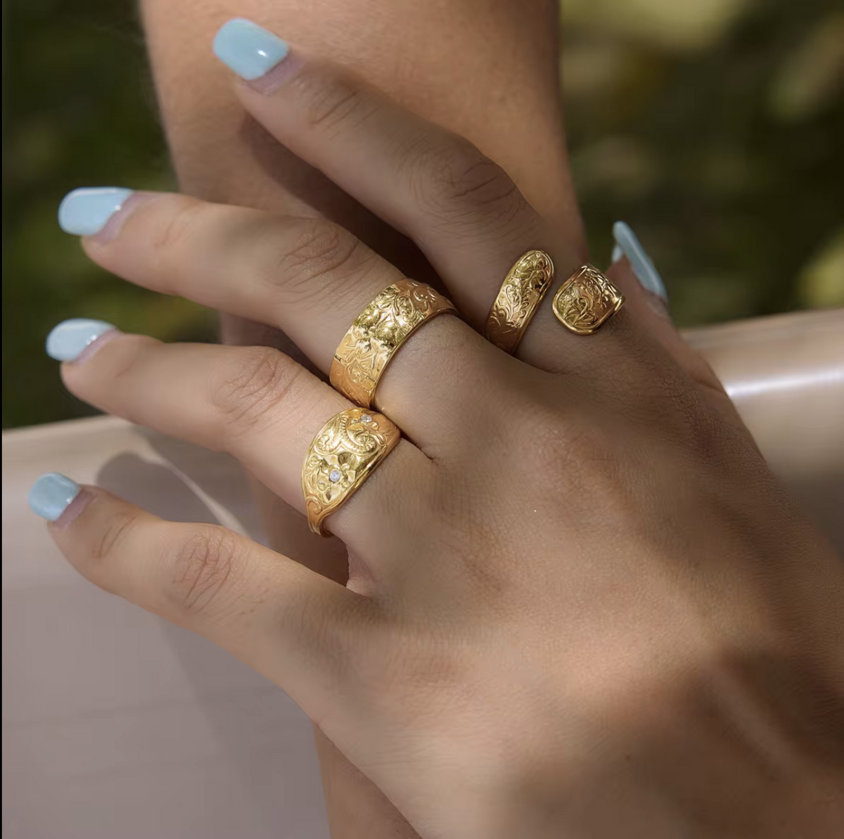 A close-up of a person's hands wearing three textured gold rings with small clear stones, set against a softly blurred outdoor background.