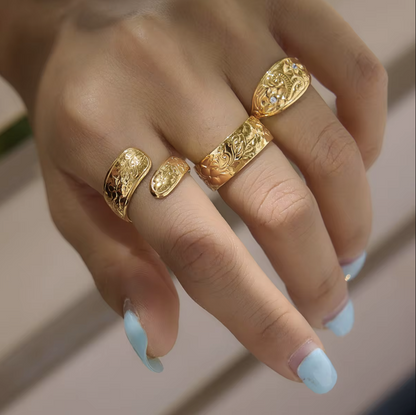 A close-up of a hand wearing four ornate gold rings with detailed floral engravings and small clear stone accents, set against a softly blurred neutral background.