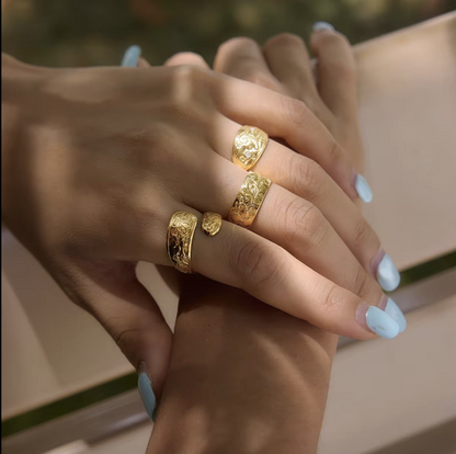 A close-up of a person's hands wearing four textured gold rings, with light blue painted fingernails and a soft-focus beige background.