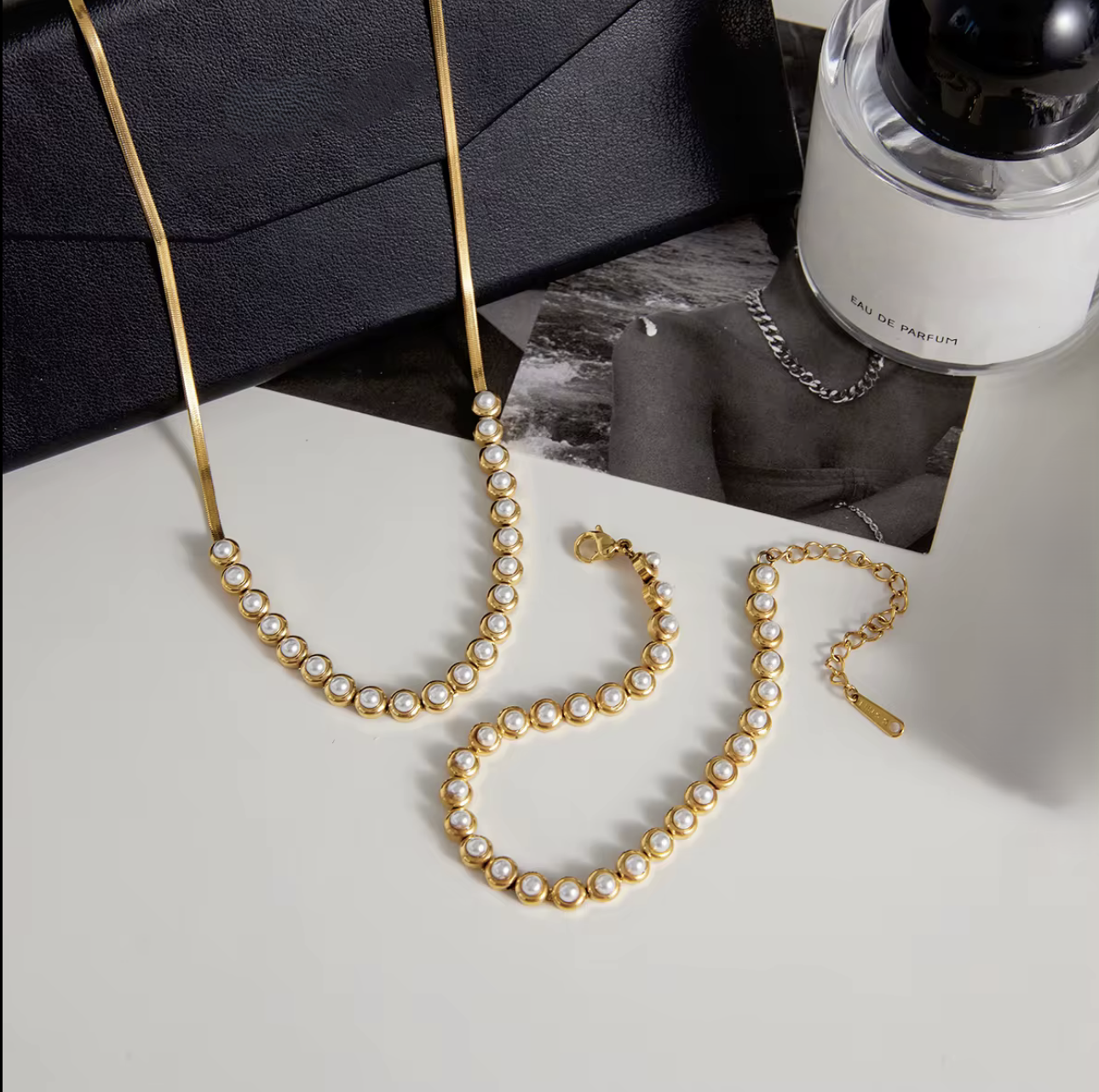 Gold necklace with pearls on a light gray surface, with a bottle of perfume in the background.