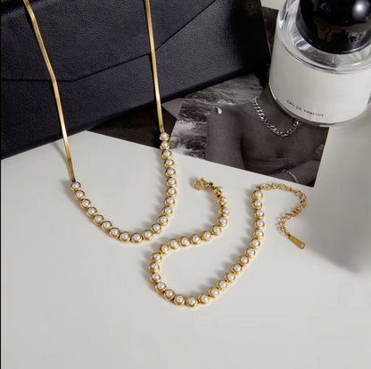 Gold necklace with pearls on a light gray surface, with a bottle of perfume in the background.
