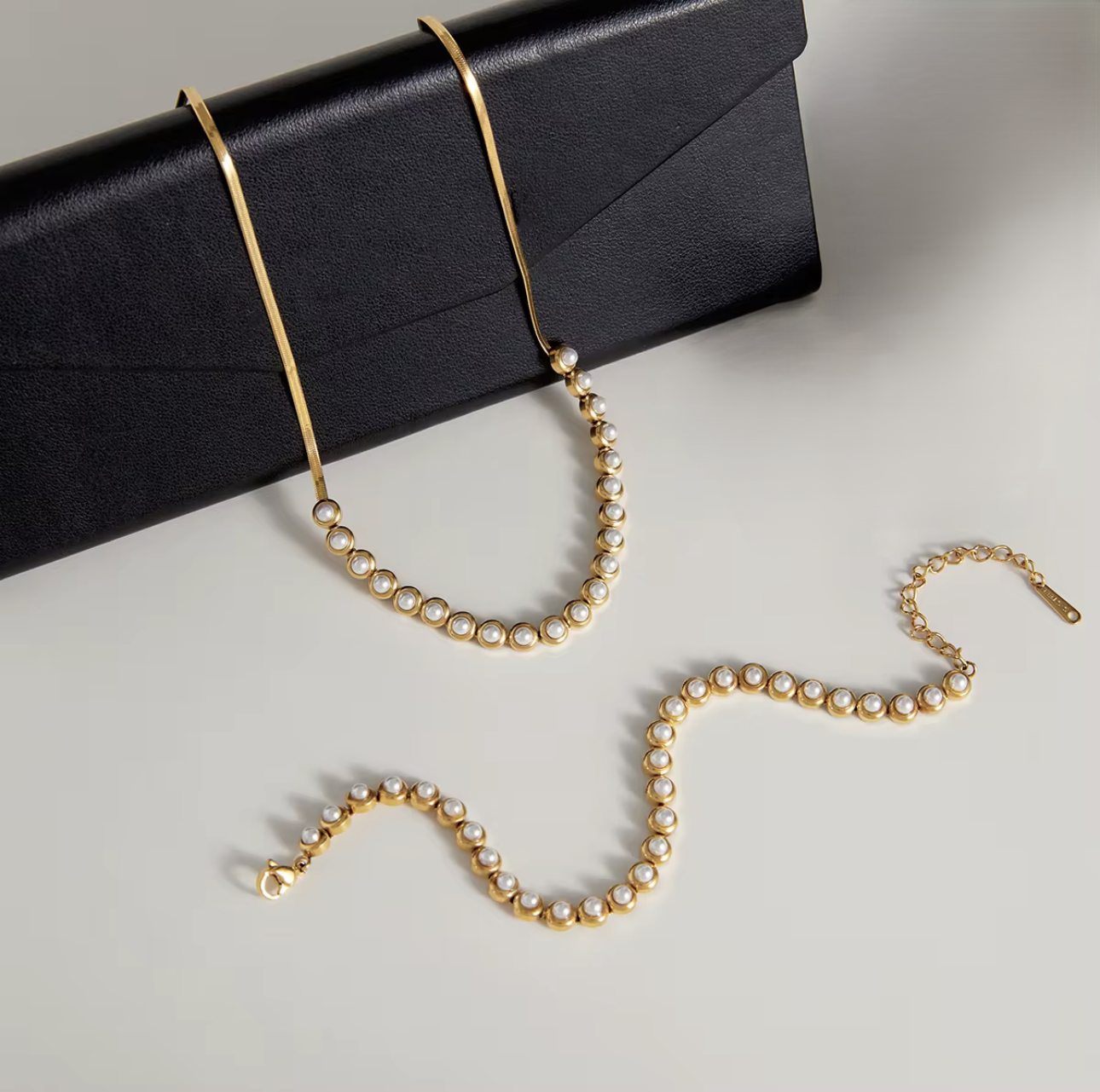 Gold necklace with pearls draped over a black box and a matching bracelet on a light gray surface