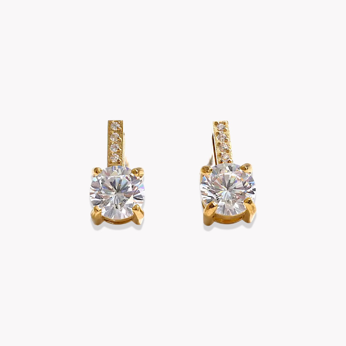 A pair of gold stud earrings featuring a large round-cut clear gemstone in a four-prong setting, with a small row of pavé-set clear stones along the top bar, shown on a light gray background.
