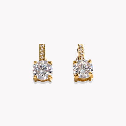 A pair of gold stud earrings featuring a large round-cut clear gemstone in a four-prong setting, with a small row of pavé-set clear stones along the top bar, shown on a light gray background.