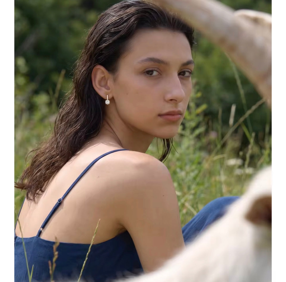 A woman outdoors wearing a navy blue spaghetti-strap top and blue jeans, looking back over her shoulder. She is wearing a small white pearl stud earring.