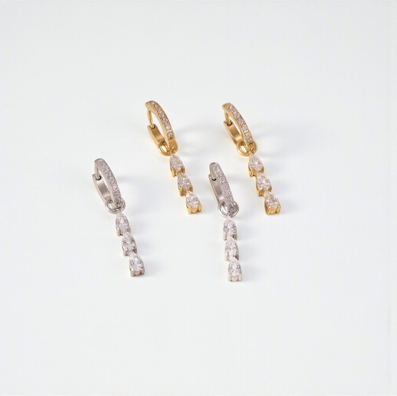 Two pairs of hoop earrings, one silver and one gold. Both with gemstone detail on the hoop and a 3 pear shaped clear gemstone drop detail. Pictured on a light grey background