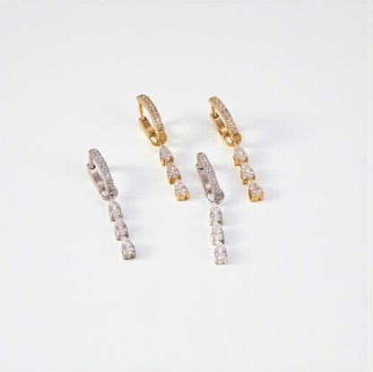 Two pairs of hoop earrings, one silver and one gold. Both with gemstone detail on the hoop and a 3 pear shaped clear gemstone drop detail. Pictured on a light grey background