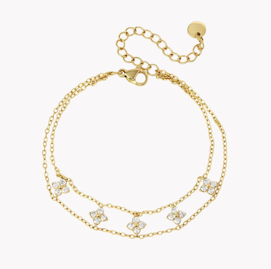 A gold double-strand chain braceletfeaturing five small four-leaf clover motifs paved with clear crystals, shown on a light gray background.