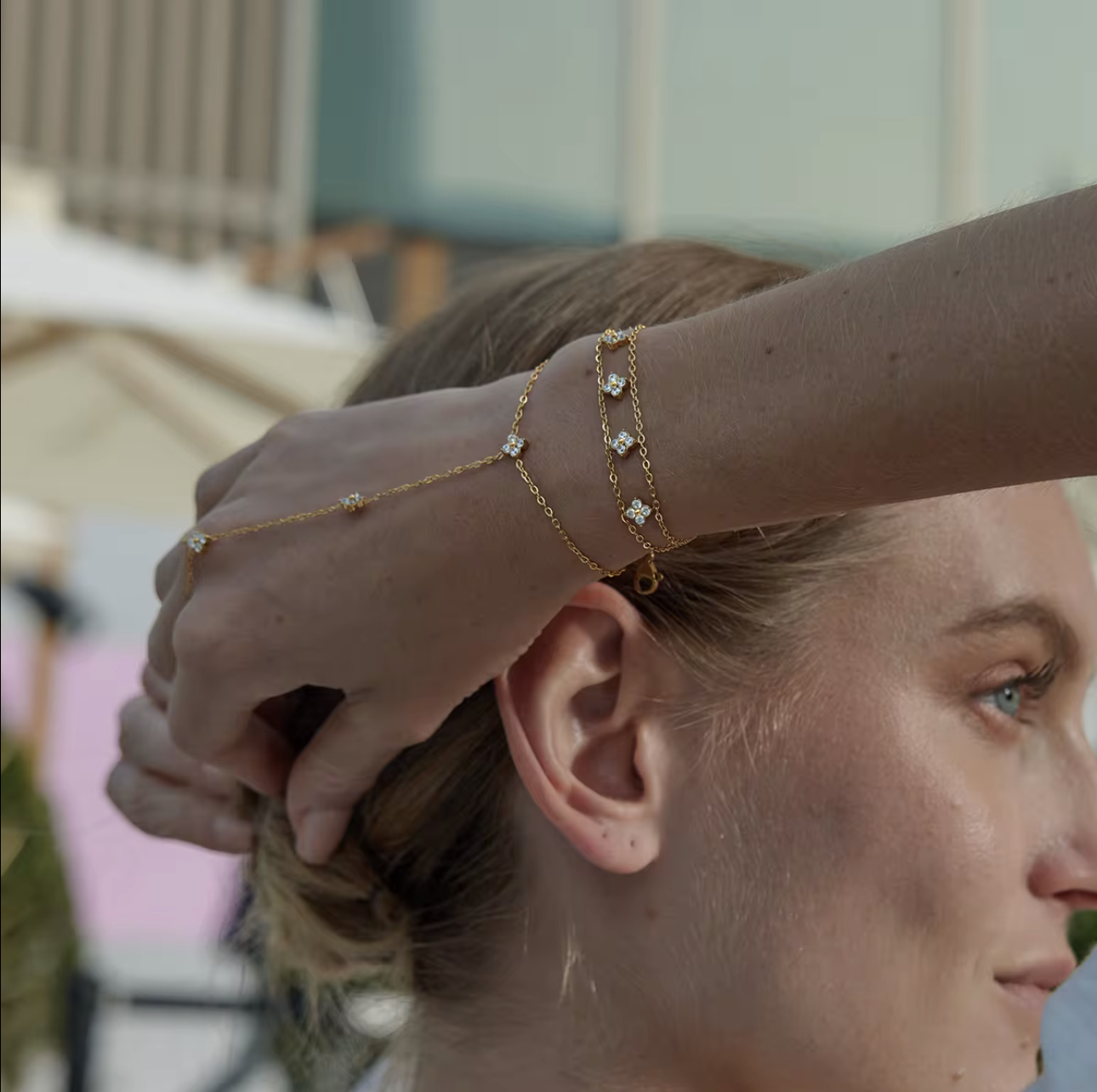 A close-up lifestyle shot of a model's hand and ear wearing delicate gold chain jewelry, including a thin chain draped over the ear and two stacked bracelets on the wrist with small clear stones.
