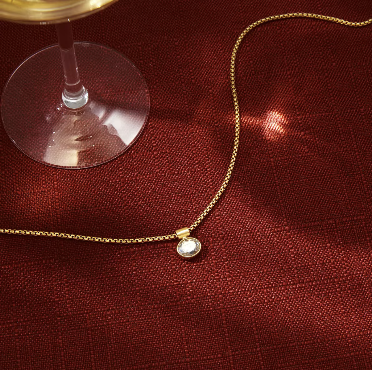 Gold necklace with a diamond pendant on a red fabric surface next to a glass of wine.