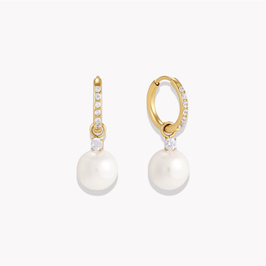 A pair of gold huggie hoop earrings adorned with small clear stones and a large white pearl drop, shown on a light gray background.