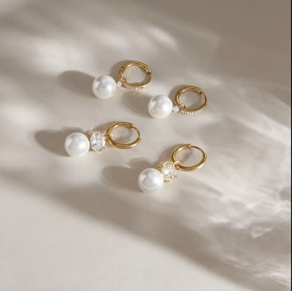 Four gold hoop earrings with white faux pearls and clear crystal accents, arranged on a warm beige surface with soft, artistic shadows.