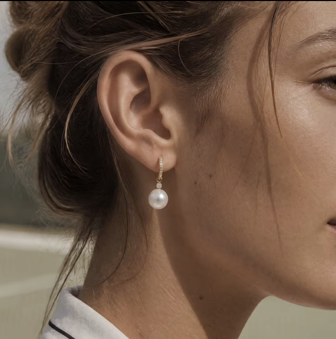A close-up of a model's ear wearing a gold huggie hoop earring adorned with small clear stones and a dangling white pearl.