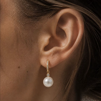 A close-up of a model's ear wearing a gold huggie hoop earring adorned with small clear stones and a dangling white pearl.