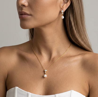 A close-up of a woman wearing a delicate gold necklace with a double pearl drop pendant and matching drop earrings, styled with a white strapless top against a neutral gray background.