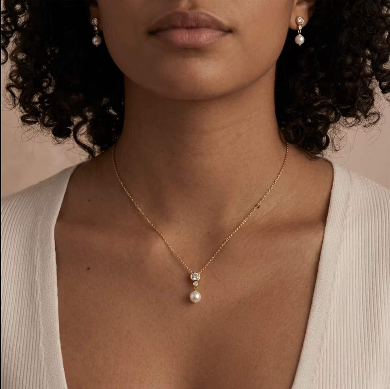 A close-up of a model wearing a delicate gold chain necklace with a white pearl pendant and matching pearl drop earrings, styled with a white ribbed top against a soft beige background.