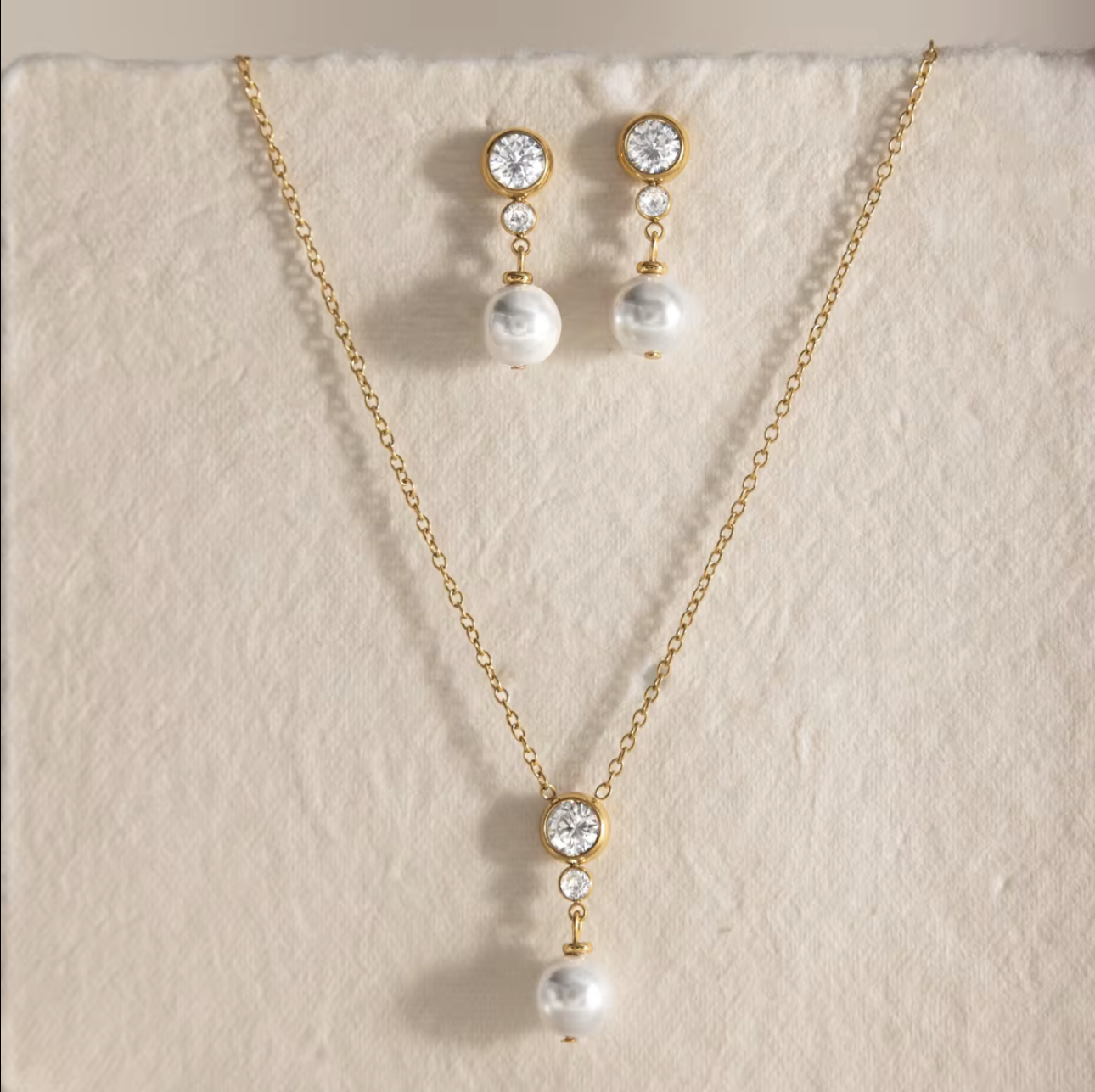 Gold-tone necklace and matching drop earrings with clear round stones and white pearl drops, displayed on a textured cream fabric background.