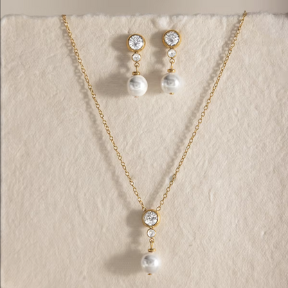 Gold-tone necklace and matching drop earrings with clear round stones and white pearl drops, displayed on a textured cream fabric background.