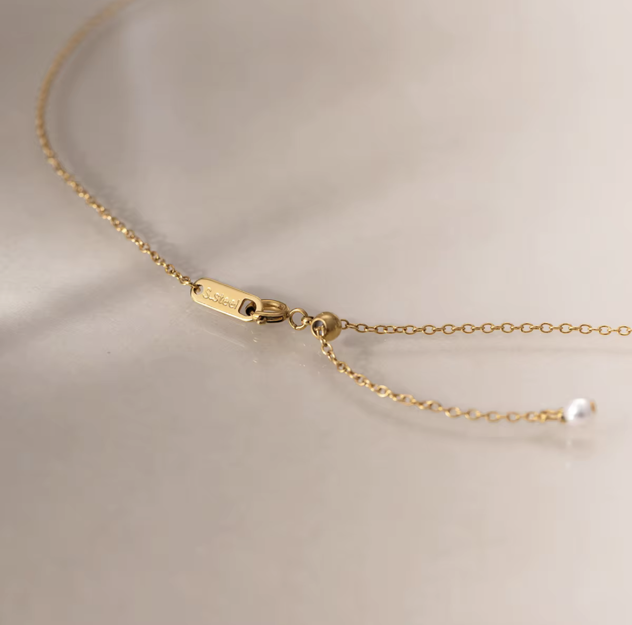 Close-up of a delicate gold chain necklace with an adjustable lobster clasp and a small white pearl bead, photographed on a soft light beige background.