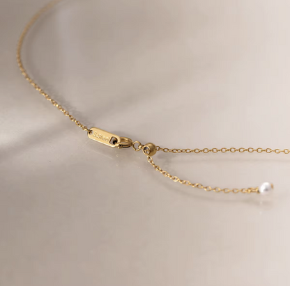 Close-up of a delicate gold chain necklace with an adjustable lobster clasp and a small white pearl bead, photographed on a soft light beige background.