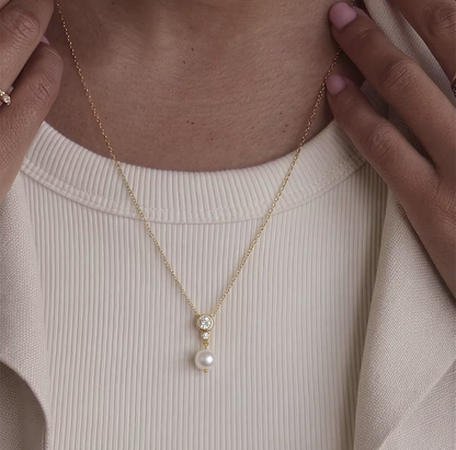 A close-up of a model wearing a delicate gold chain necklace with a small clear stone and a dangling white pearl, styled over a cream ribbed top.